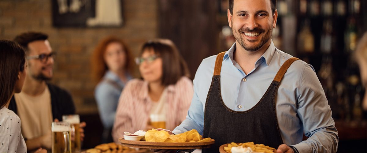 Happy waiter holding plates with food and looking at camera while serving guests in a restaurant.